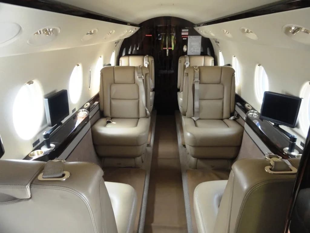 The interior cabin of a private jet with light brown leather seats arranged in a club configuration, and dark wood accents.