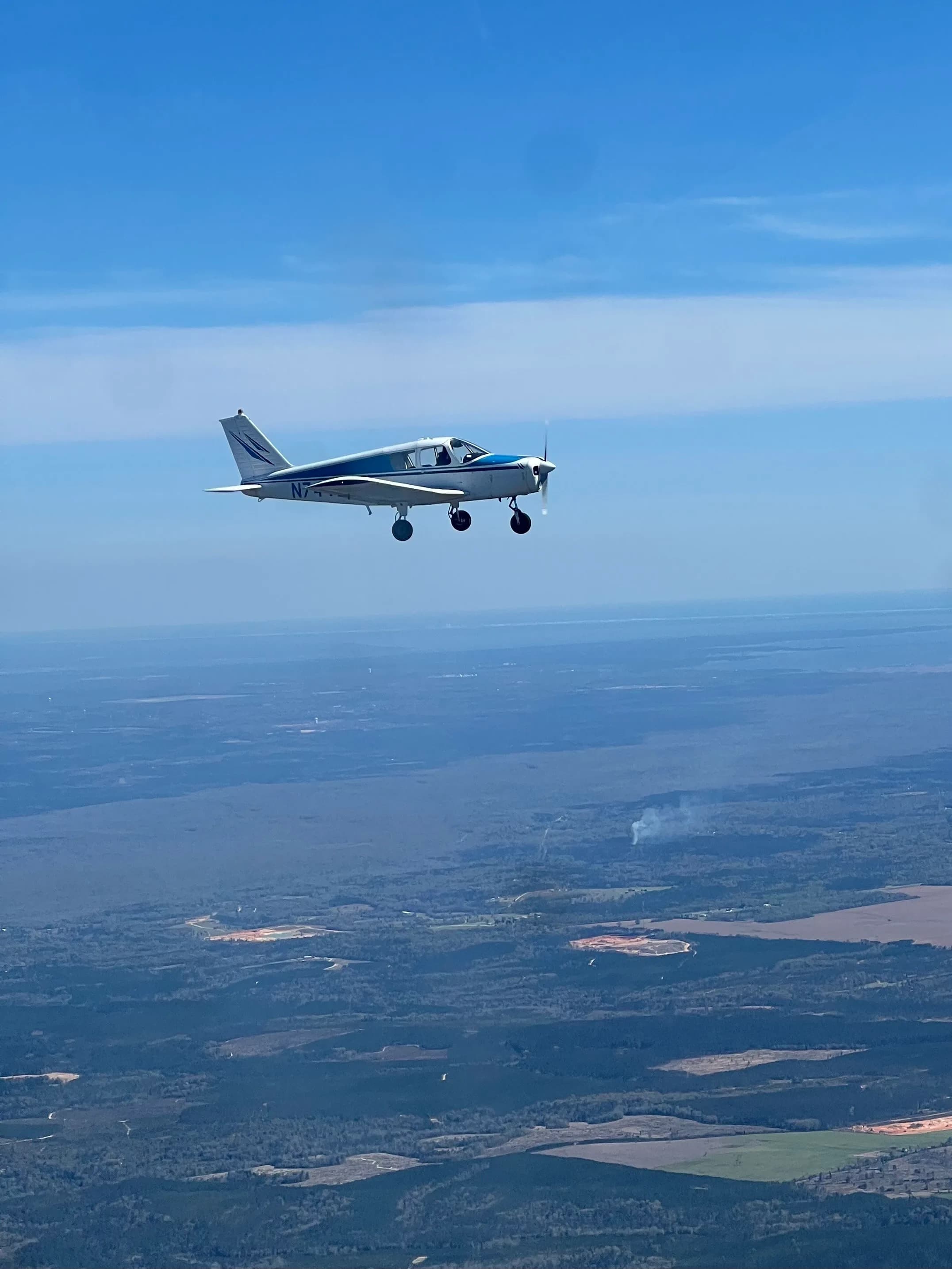 A white and blue Piper Cherokee 140 (N7443R) flying over a landscape with forests and fields.
