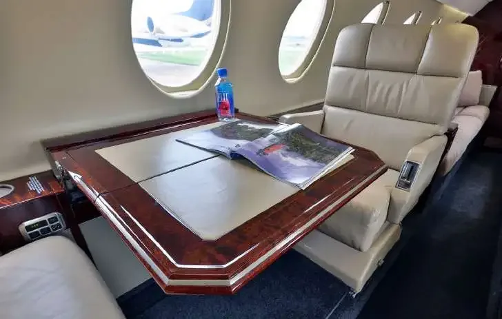 A beige leather seat next to a polished wooden table with a magazine and water bottle, looking out an aircraft window.