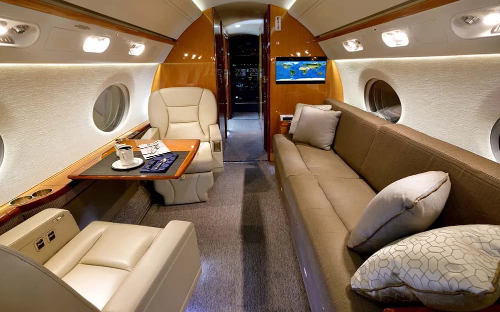 A section of the aircraft cabin with a long brown sofa, a beige seat, and a table set with coffee and a newspaper.