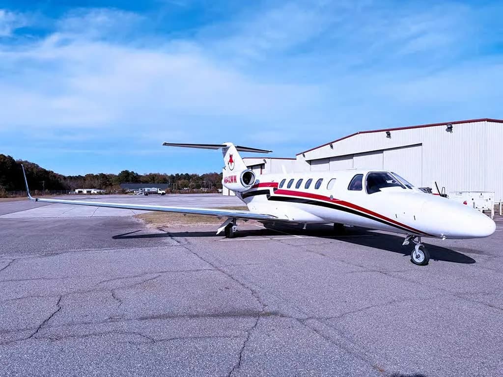 A white Cessna Citation private jet with red and black stripes and tail logo, parked on an asphalt tarmac with buildings and trees in the background.