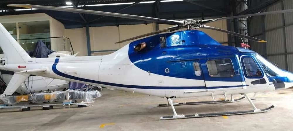 Side view of a blue and white helicopter with its main rotor blades extended, parked inside a hangar, with another aircraft in the background.