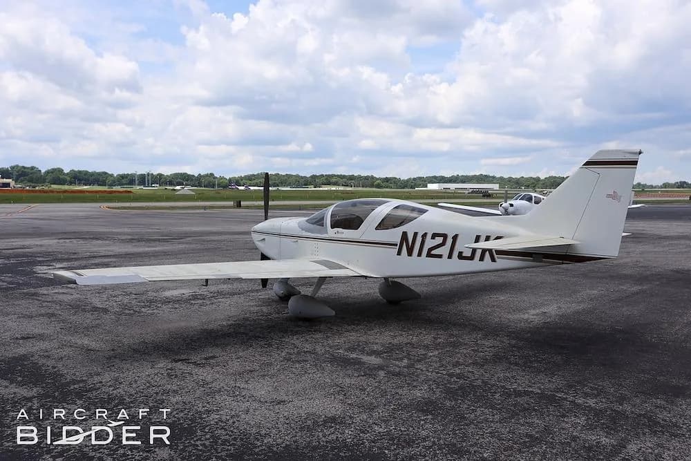 A white single-engine aircraft (N121JK) with brown stripes parked on an asphalt apron at an airfield.
