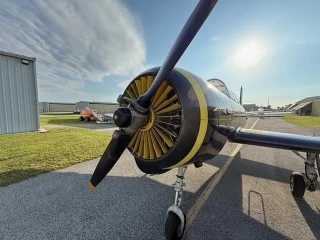 A close-up, low-angle view of the propeller and engine cowling of a dark gray Yak-52 aircraft with a yellow spinner and engine fins.