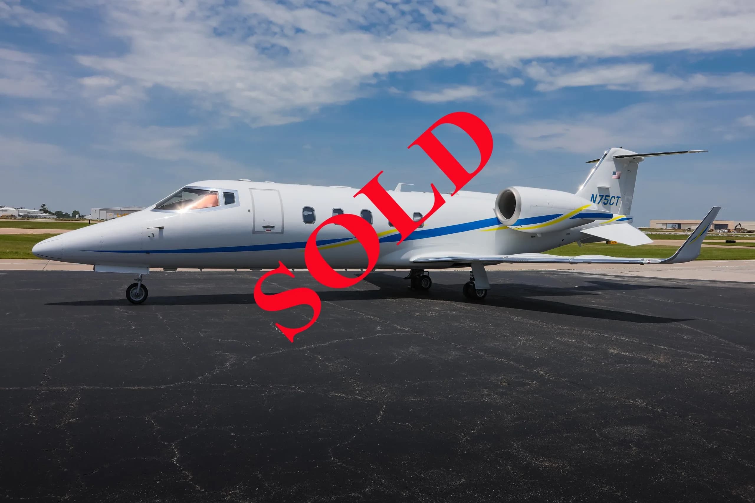 A white business jet with blue and yellow stripes, registration N75CT, parked on an asphalt tarmac under a partly cloudy sky.