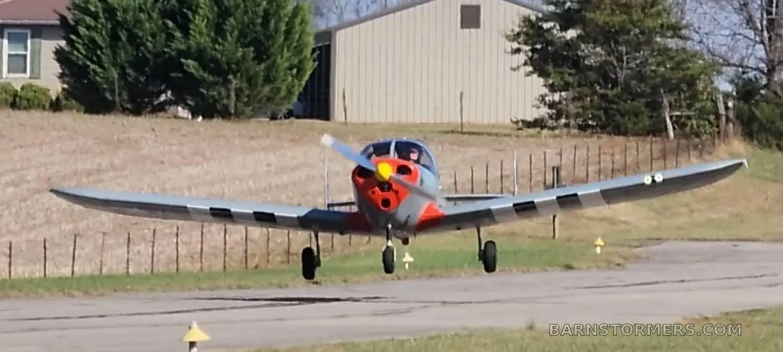 A grey single-engine aircraft with a red nose is seen from the front, just above a grassy airstrip.