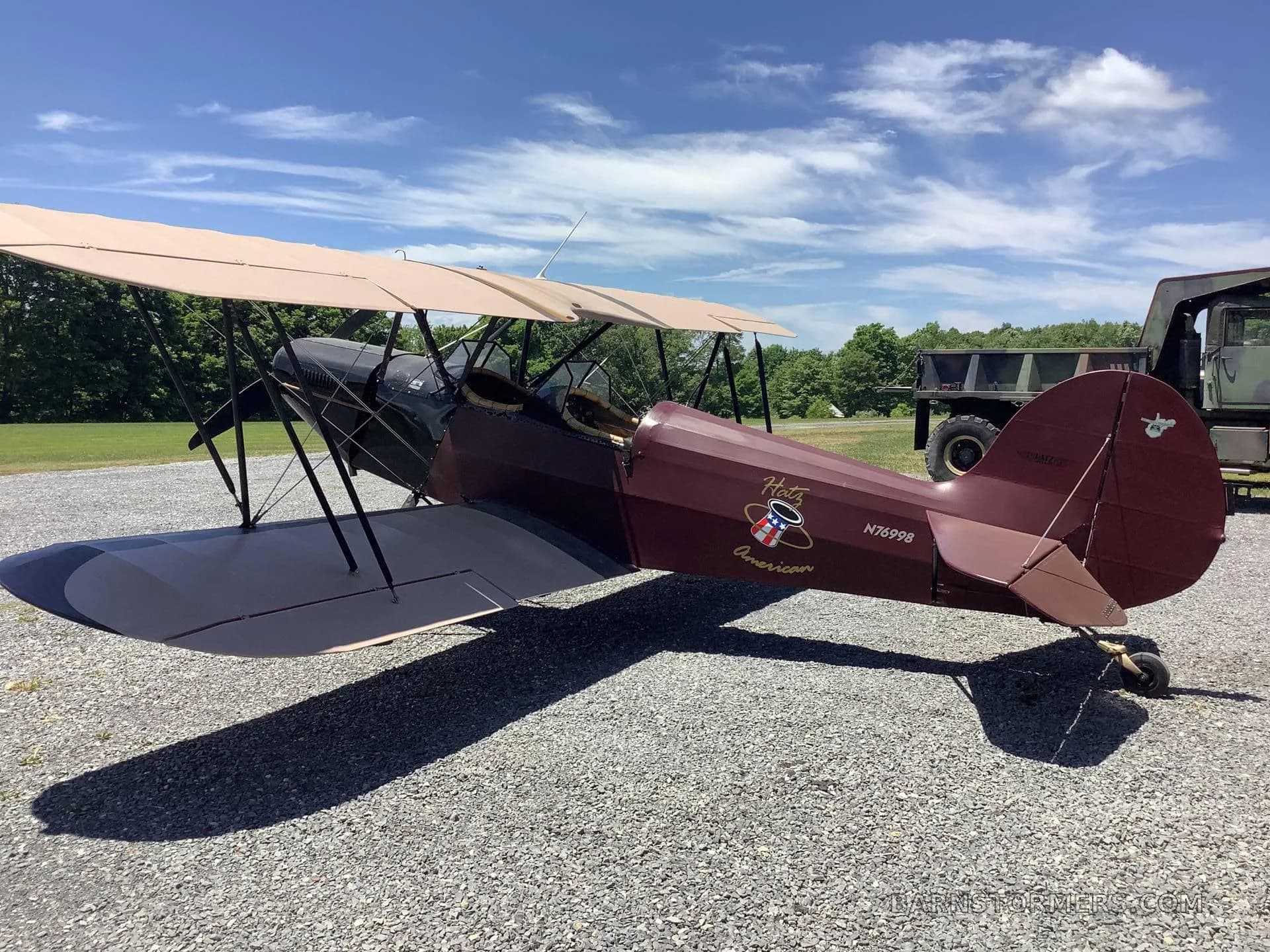 A maroon and black biplane with a 'Hotz American' logo, parked on a gravel path with a military truck in the background.