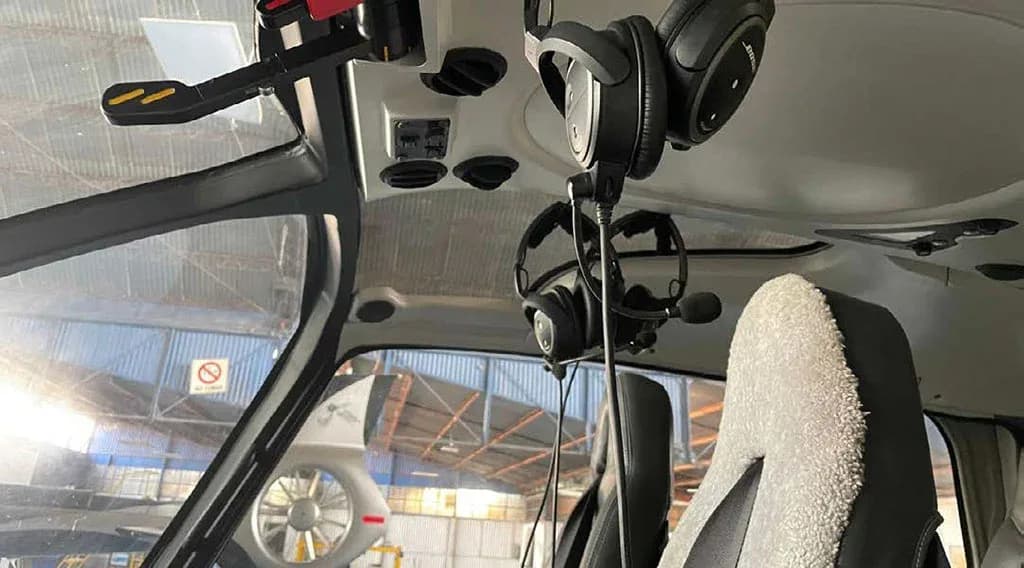 Headsets hang in the front cabin of the helicopter, with a view into a hangar through the window.