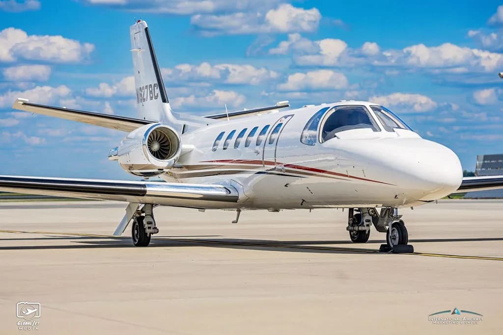 A white Cessna Citation Bravo business jet with red and black stripes on the tarmac under a blue sky.