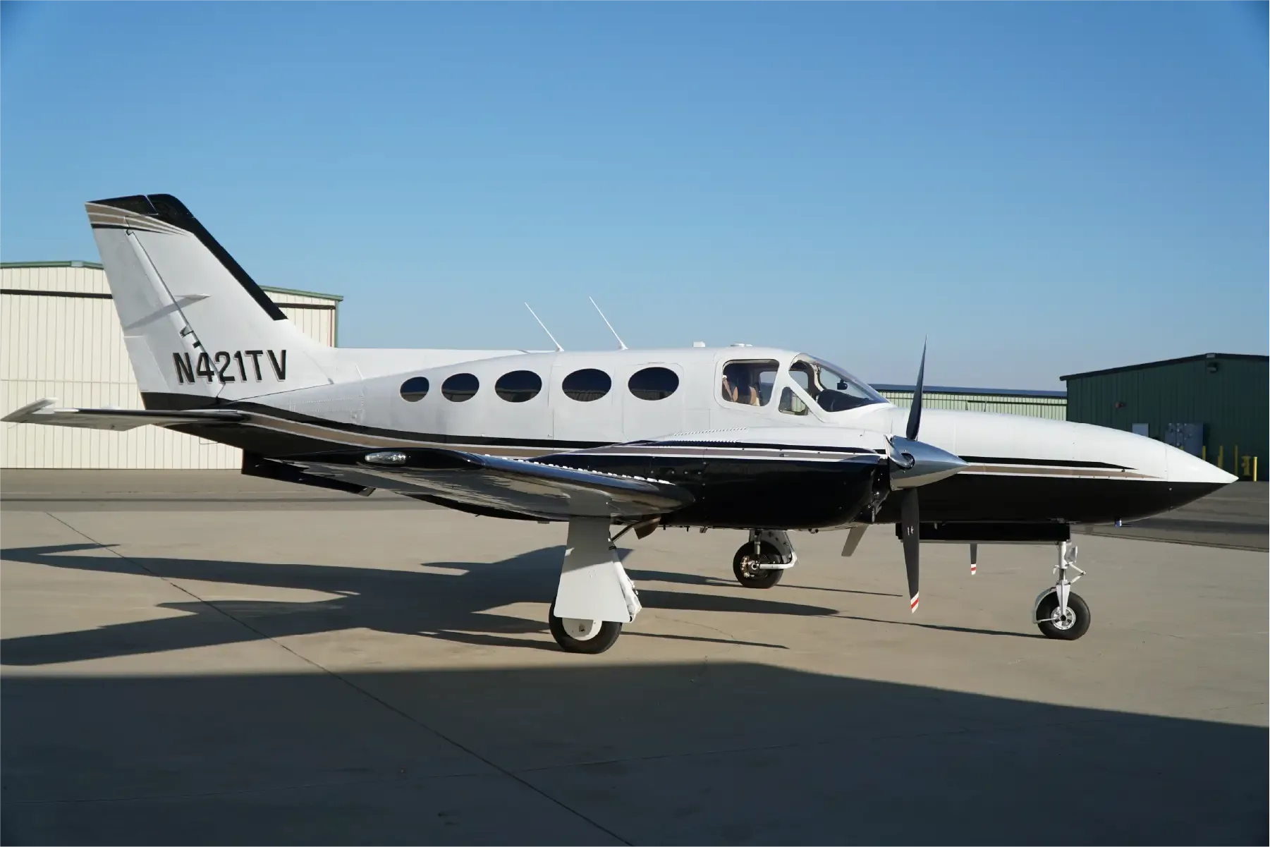 A white and black twin-engine propeller aircraft, likely a Cessna 421, parked on an airfield with hangars in the background under a clear blue sky.