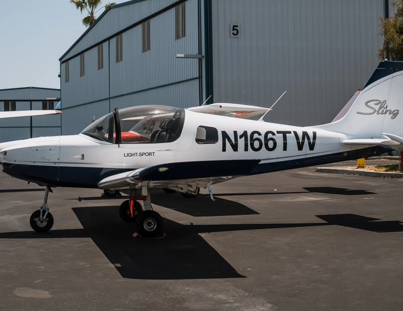 A white and blue Sling Light-Sport aircraft, N166TW, parked on an asphalt apron next to a hangar.