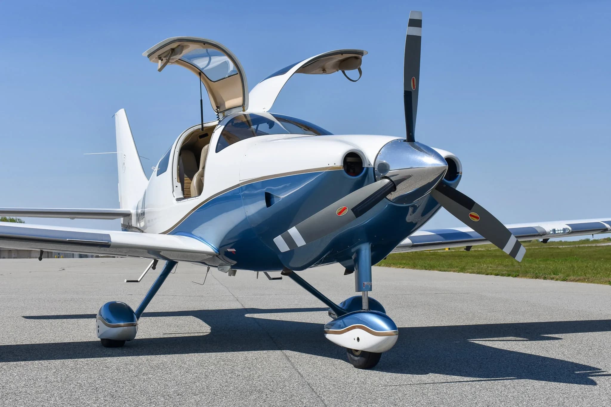 Front view of a white and blue single-engine aircraft with open gull-wing doors on an airfield.