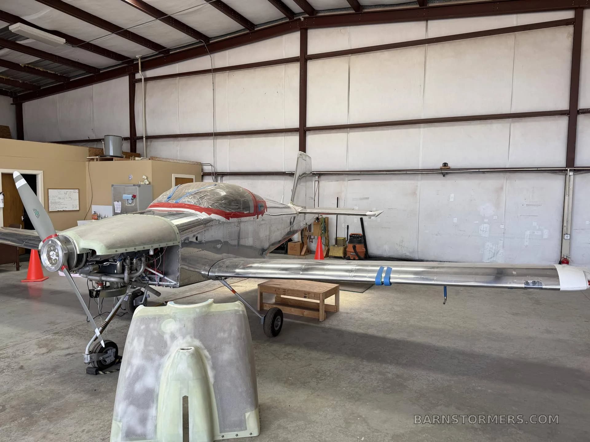 A partially assembled, unpainted metal aircraft with a red-taped canopy and exposed engine in a hangar.