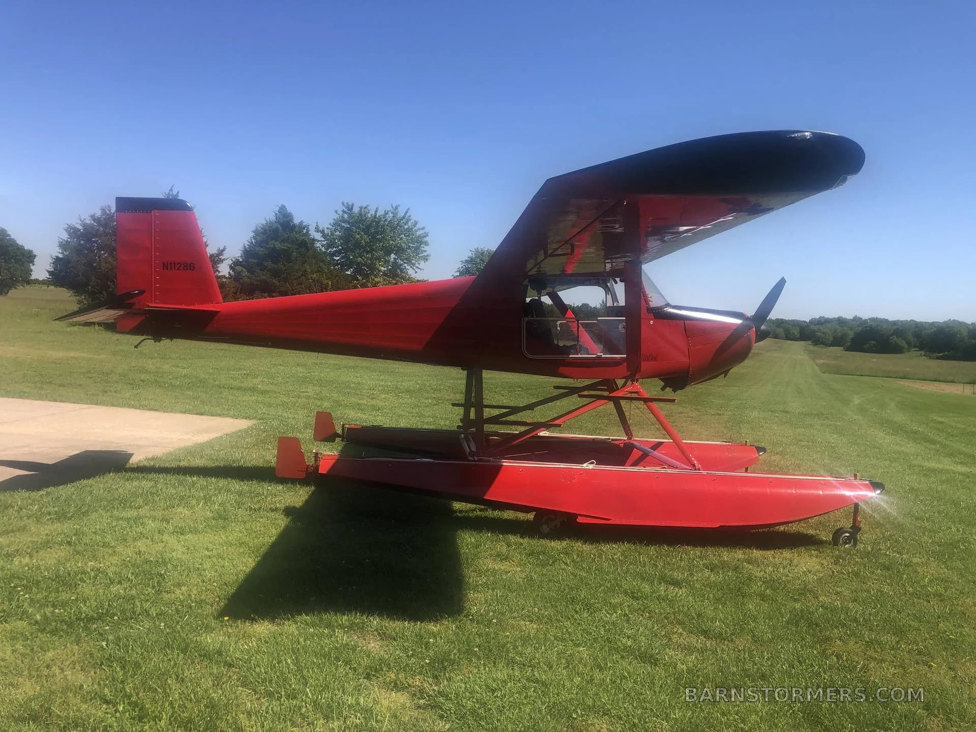 A red floatplane with registration N11286 is parked on green grass next to a concrete pad under a clear blue sky.