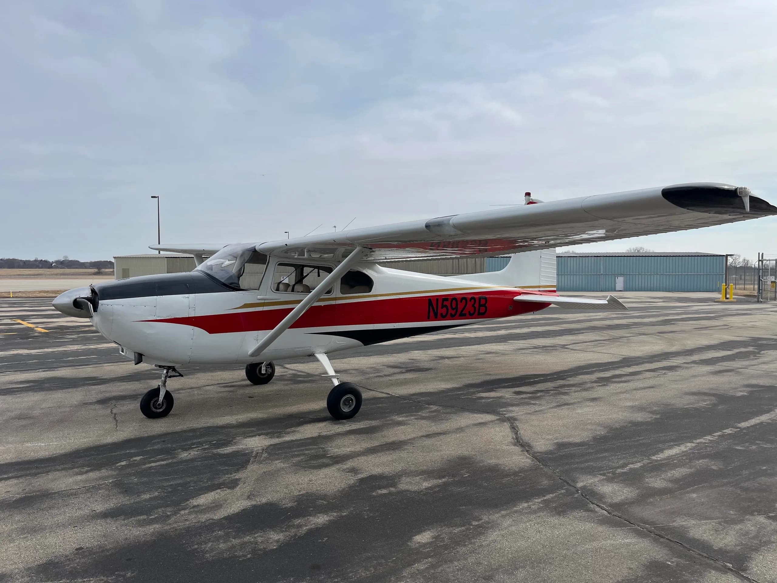 A side view of a white Cessna 182 aircraft with red and black stripes and registration N5923B, parked on an asphalt apron.