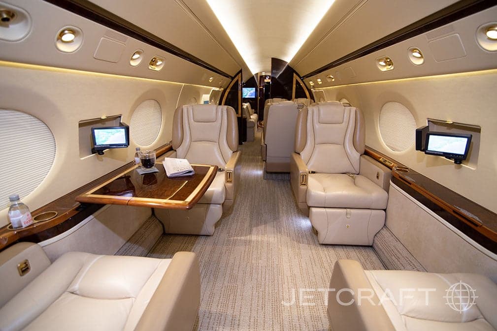 A view down the aisle of a Gulfstream G550 cabin, showing beige leather seats, a fold-out table, and individual entertainment screens.