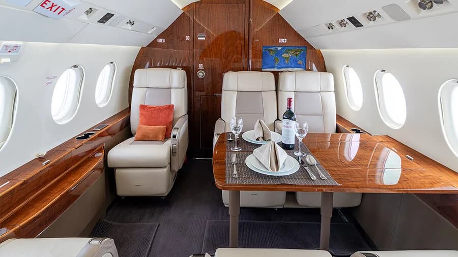 A private jet cabin section with a dining table set for two, featuring a bottle of red wine, place settings, and beige leather seats.
