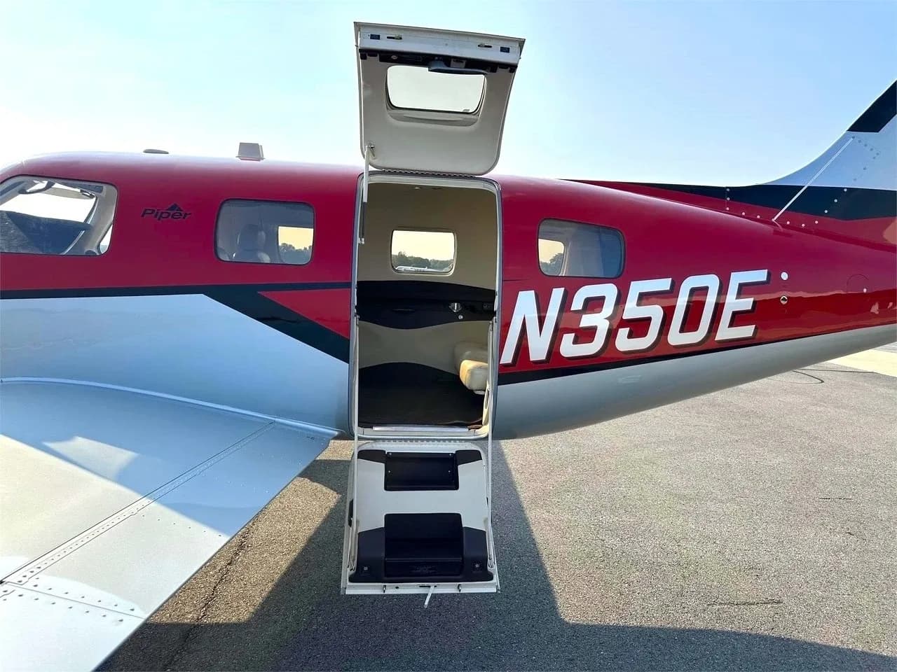 The open cabin door and airstair of a red and silver Piper M350 aircraft, registration N350E, on an airport tarmac.
