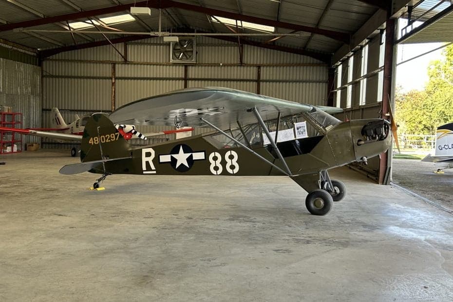 A Piper J-3 Cub aircraft is shown in a hangar with a military-style paint scheme.