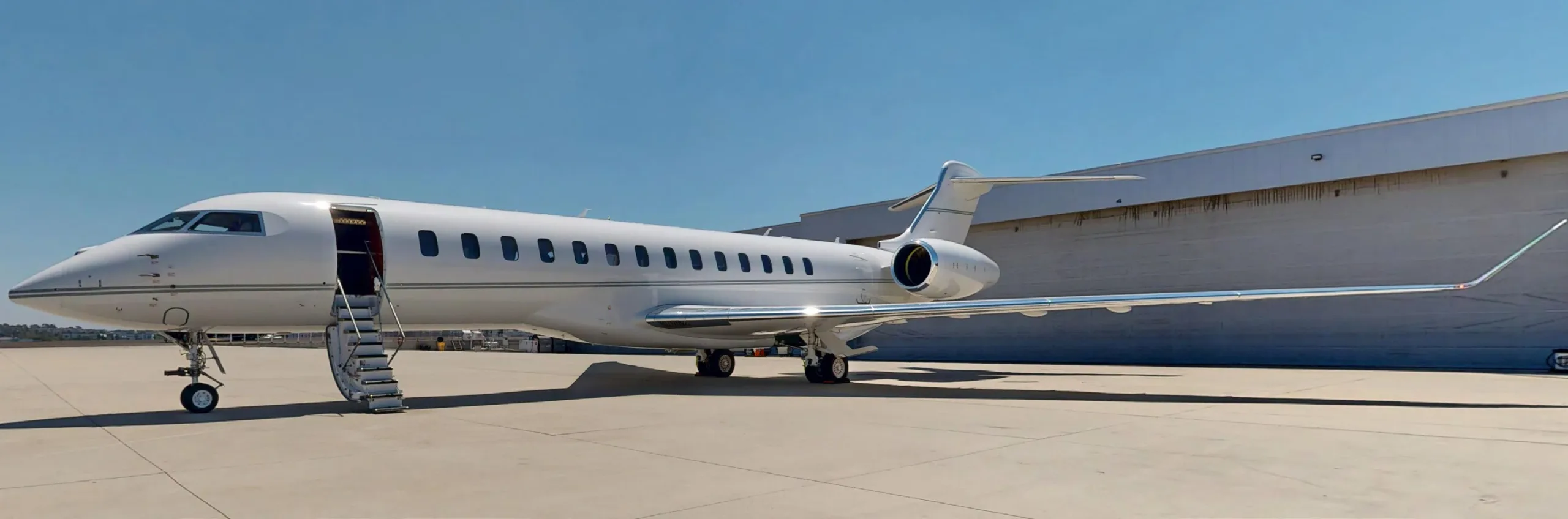 A white Bombardier Global 6000 private jet with its door open and stairs extended, parked on a tarmac next to a hangar on a sunny day.