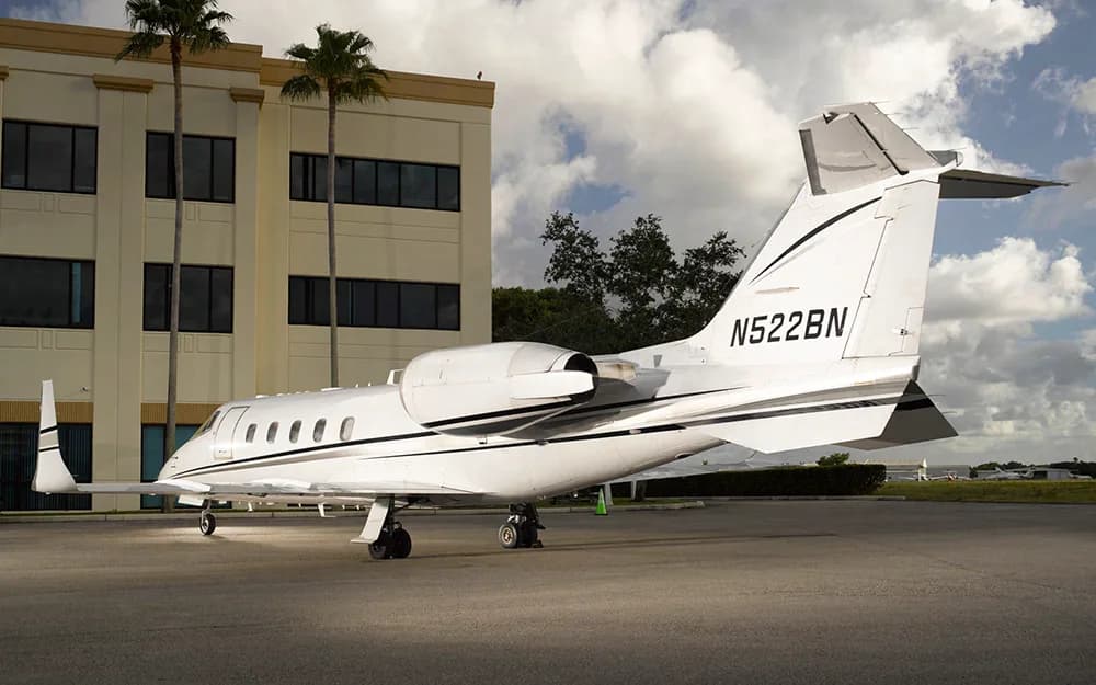 A white Learjet with black and grey stripes, tail number N522BN, parked next to a tan building with palm trees.