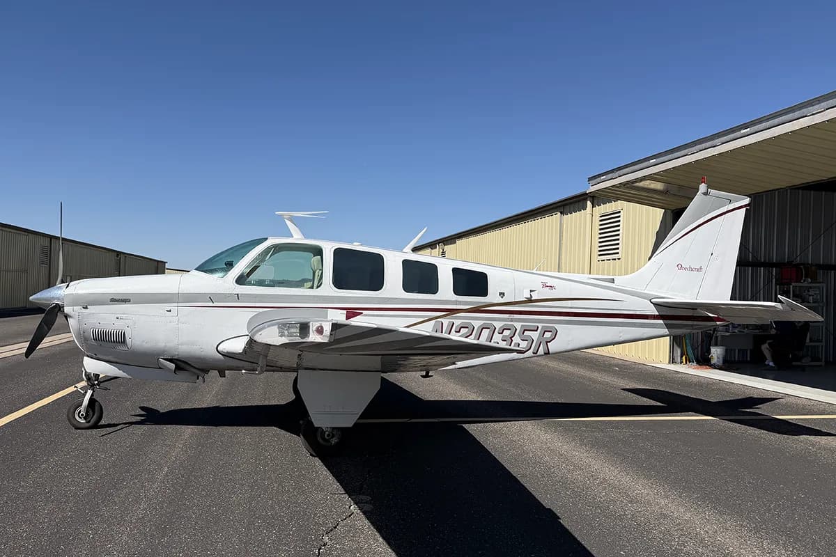 A white Beechcraft Bonanza with maroon and grey stripes is parked on an asphalt tarmac next to a hangar on a sunny day.