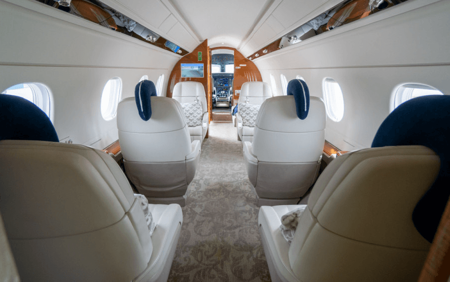 A view down the aisle of a private jet cabin, showing multiple rows of light-colored leather seats and the cockpit entrance in the distance.