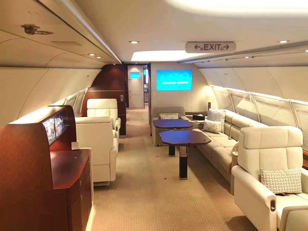 A spacious private jet interior featuring beige seating, tables, and a screen, viewed from a different angle.