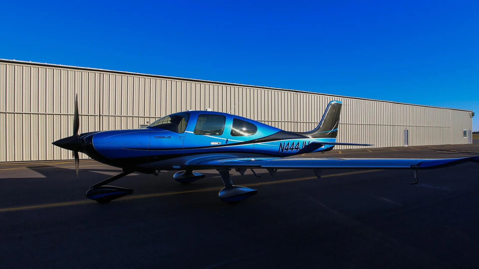 A blue and silver Cirrus SR22 aircraft with registration N444JH parked on a tarmac under a clear blue sky.