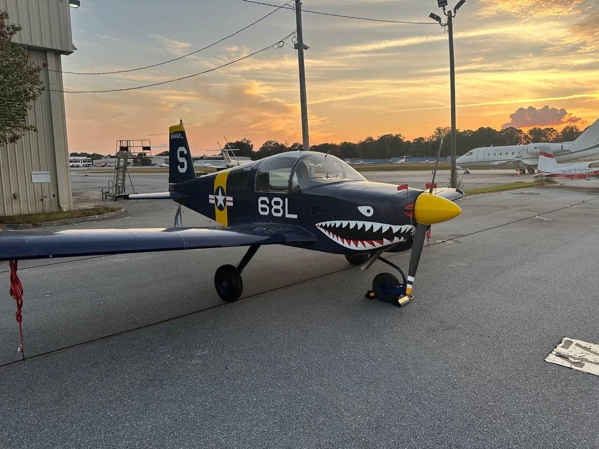 A blue and yellow aircraft with shark teeth painted on the nose is parked on the tarmac at sunset.