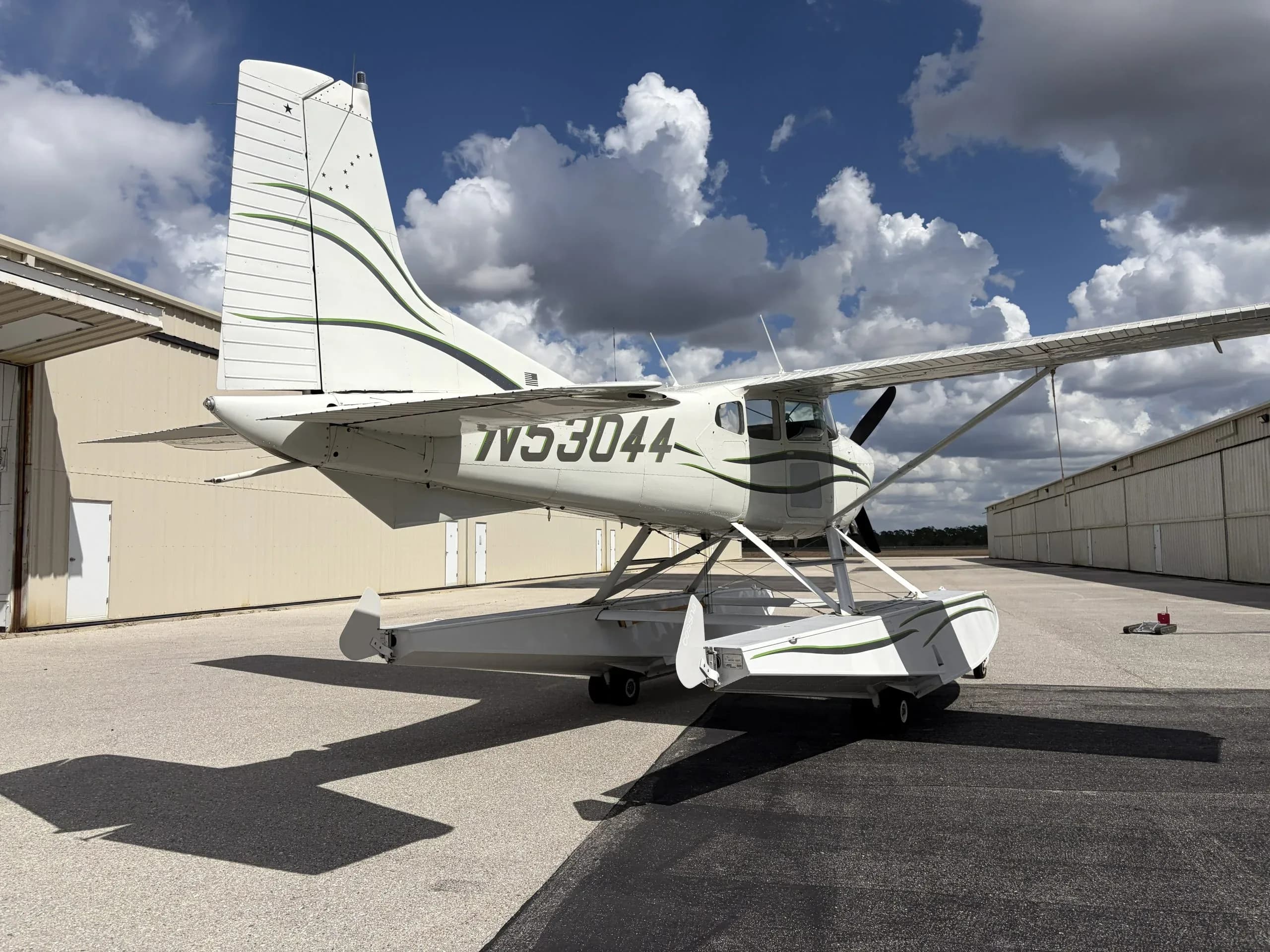 A rear-side view of a white Cessna 180 Skywagon seaplane with green stripes, registration N53044, parked on an asphalt apron.