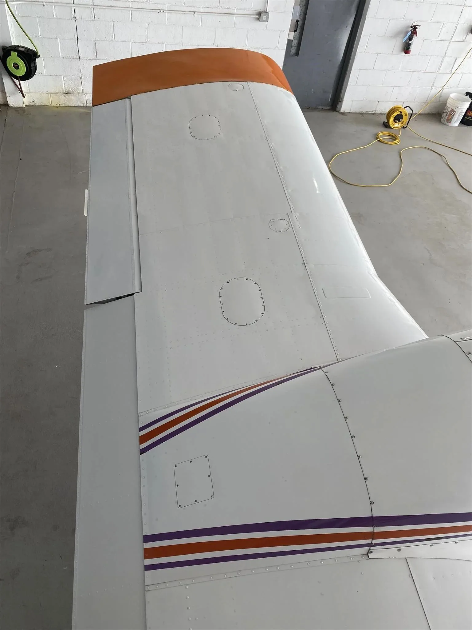 An overhead view of the right wing and fuselage of the white Piper Aztec aircraft, showing the orange wingtip and purple stripes.