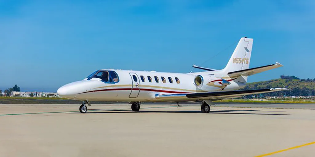 A white business jet with red and gold stripes, registration N554TS, parked on an airport tarmac under a clear blue sky.
