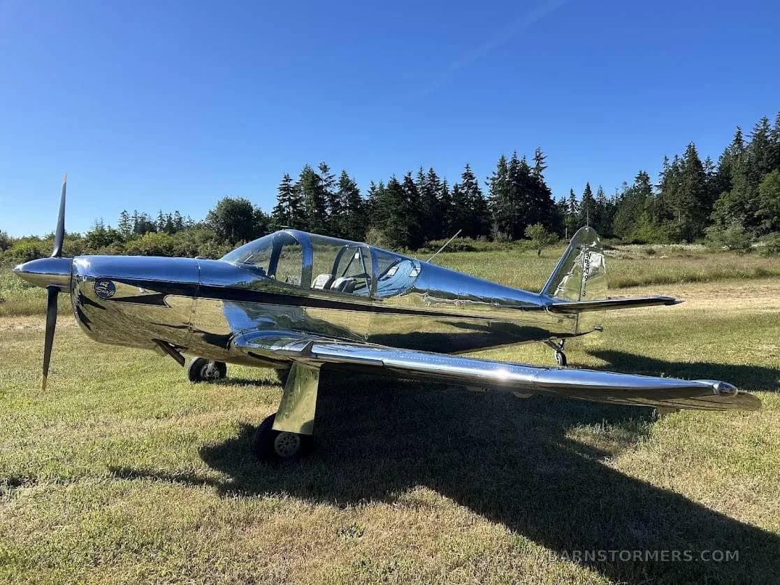 A shiny, chrome-like single-engine propeller aircraft with a bubble canopy is parked on a grassy field with trees in the background.