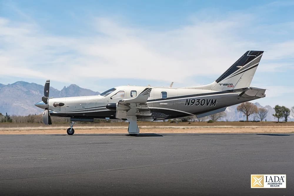A side view of the N930VM TBM 930 aircraft in white and black livery on an airfield with mountains in the background.