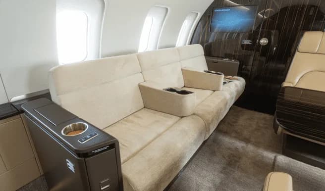 A beige sofa-style seat with cup holders and a side console inside a private jet cabin.