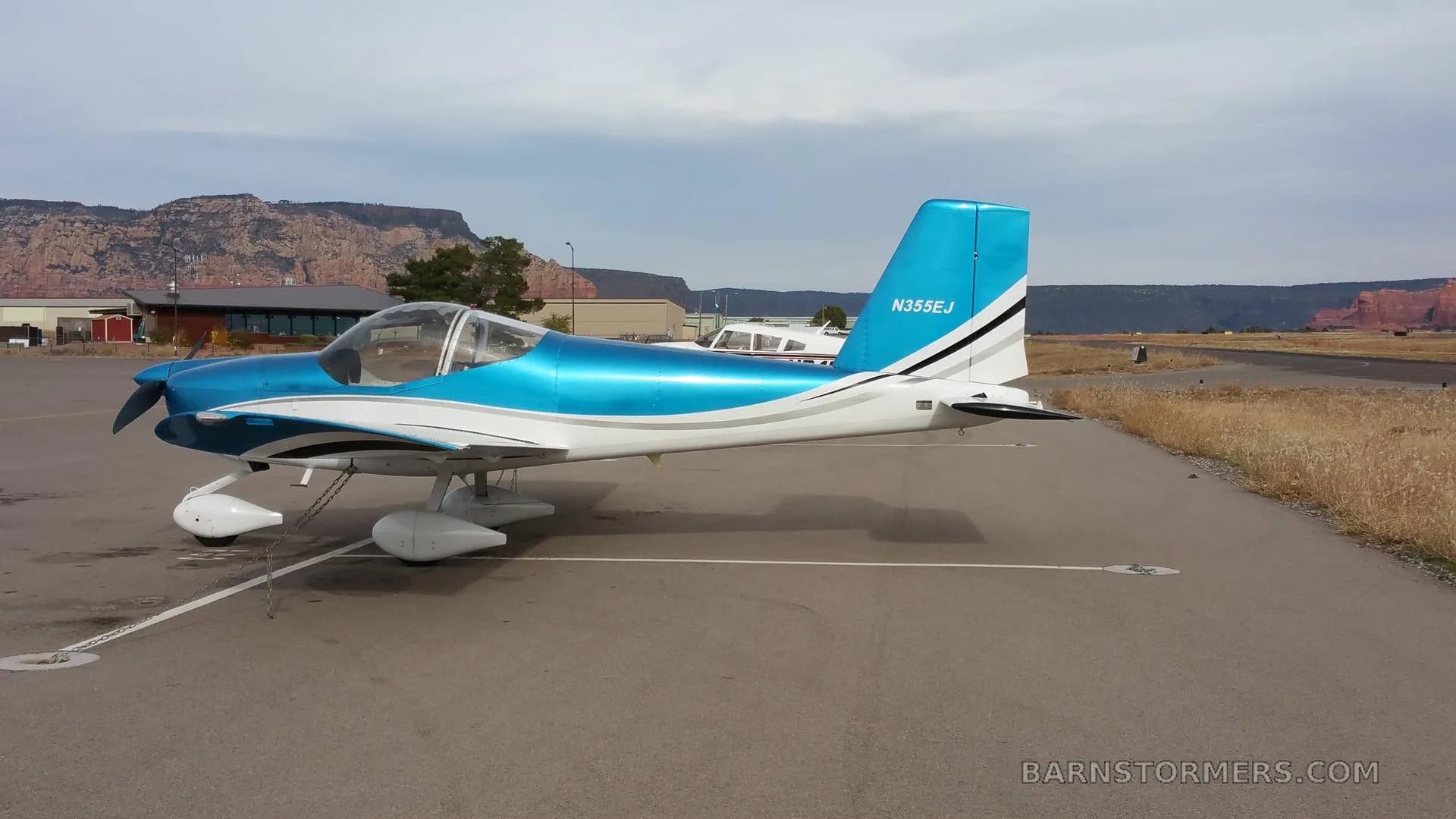 A blue and white single-engine aircraft with registration N355EJ is parked on an airfield, viewed from the side with distant mountains.