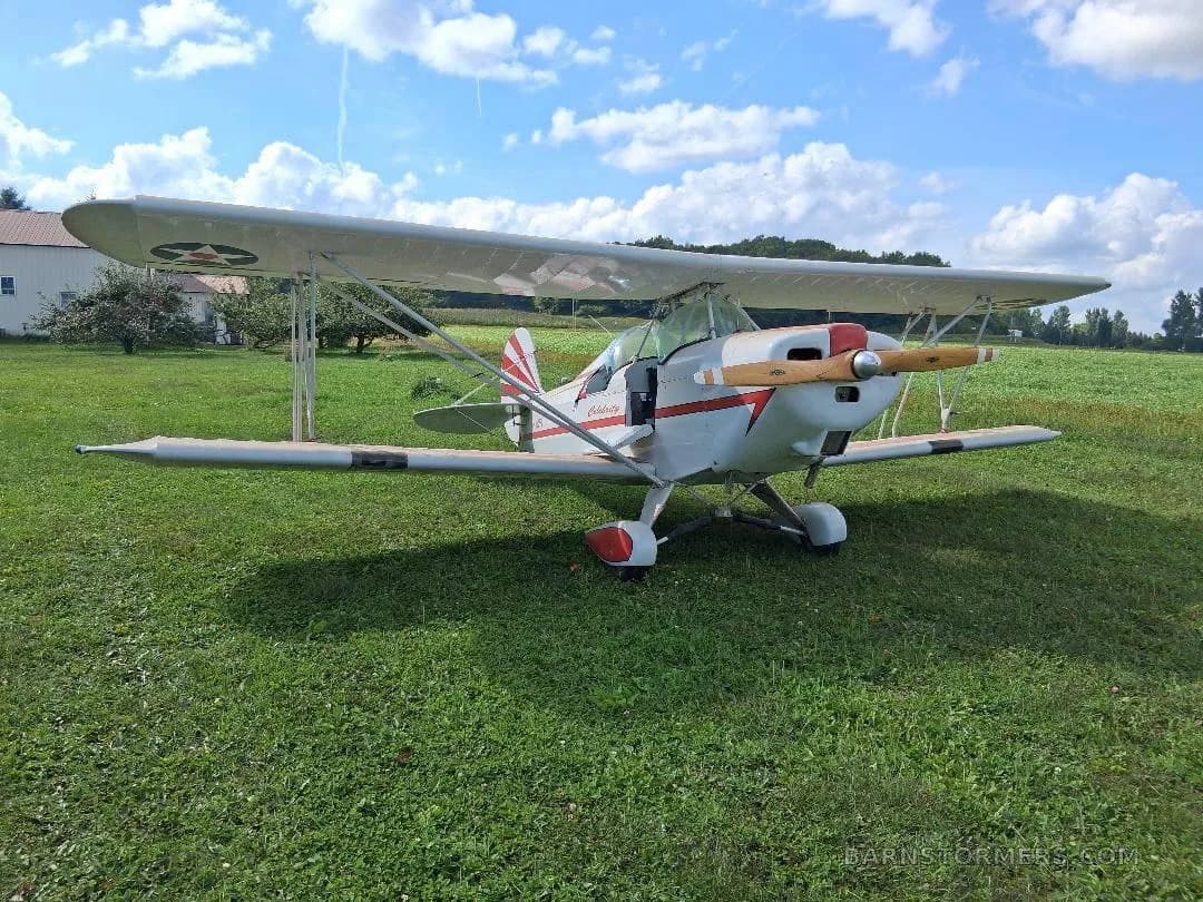A white biplane with red stripes and a wooden propeller, viewed from the front-right side, parked on green grass.