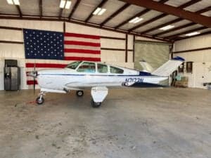 A Beechcraft Bonanza aircraft parked in a hangar with an American flag in the background.