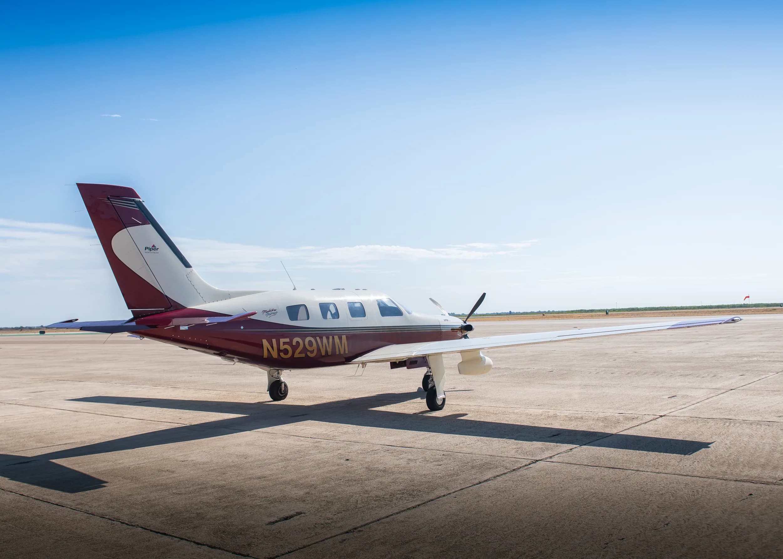 A three-quarter rear view of the maroon and white Piper M350 aircraft (N529WM) parked on an airfield under a bright blue sky.