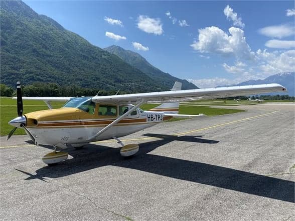 A Cessna 210 Centurion aircraft with a white and gold paint scheme is parked on the tarmac with mountains in the background.