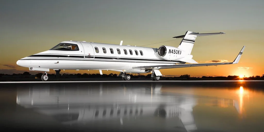 A white business jet with black stripes, registration N450KV, sits on a tarmac at sunset with reflections on the wet ground.