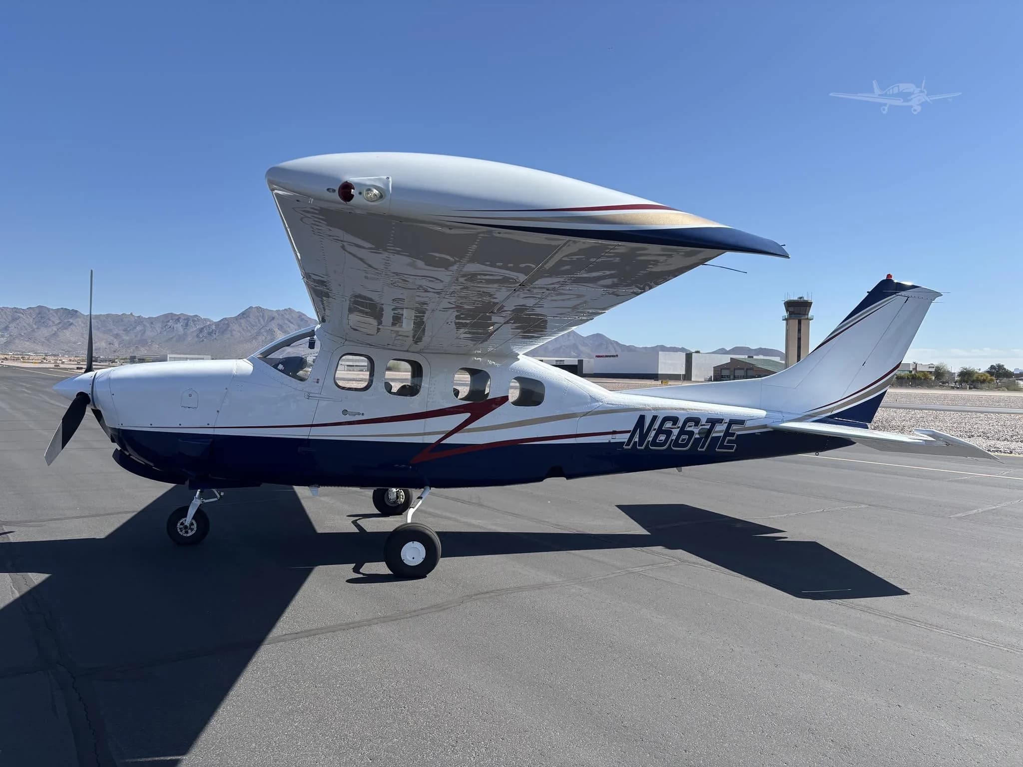 A white and blue Cessna 206 Stationair with red and gold stripes parked on an airport tarmac with mountains and a control tower in the background.