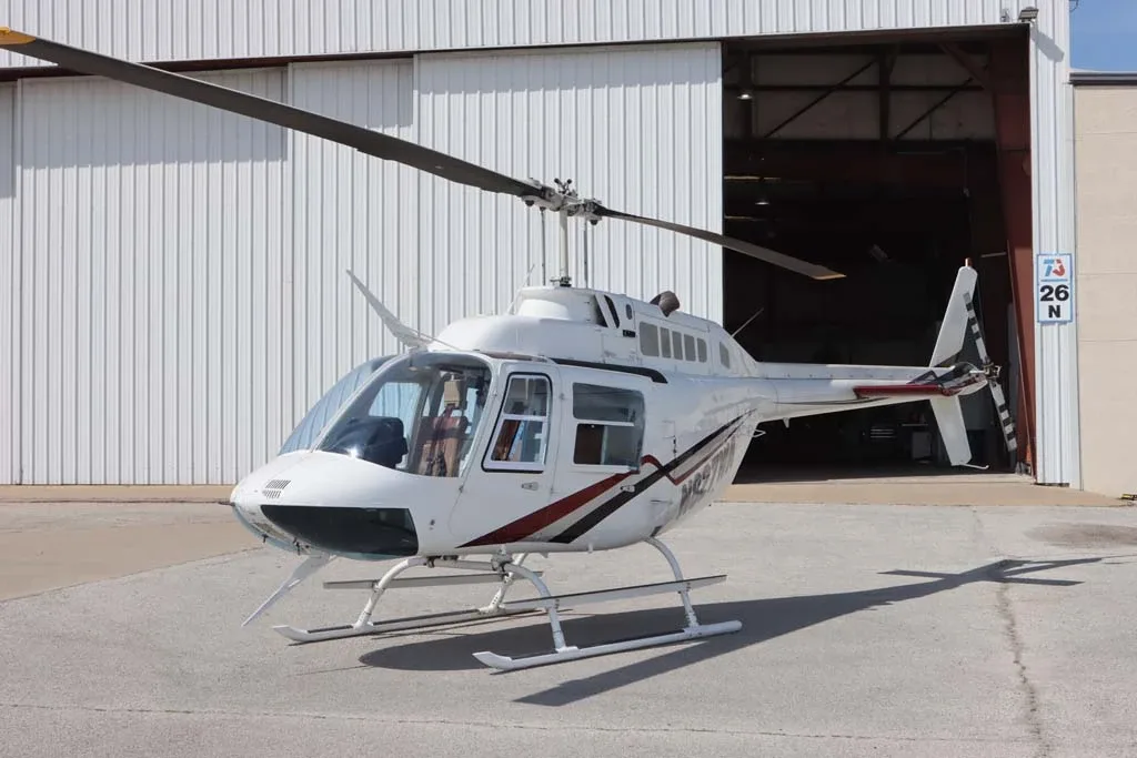 A white Bell 206 helicopter with red and black stripes parked in front of a hangar.