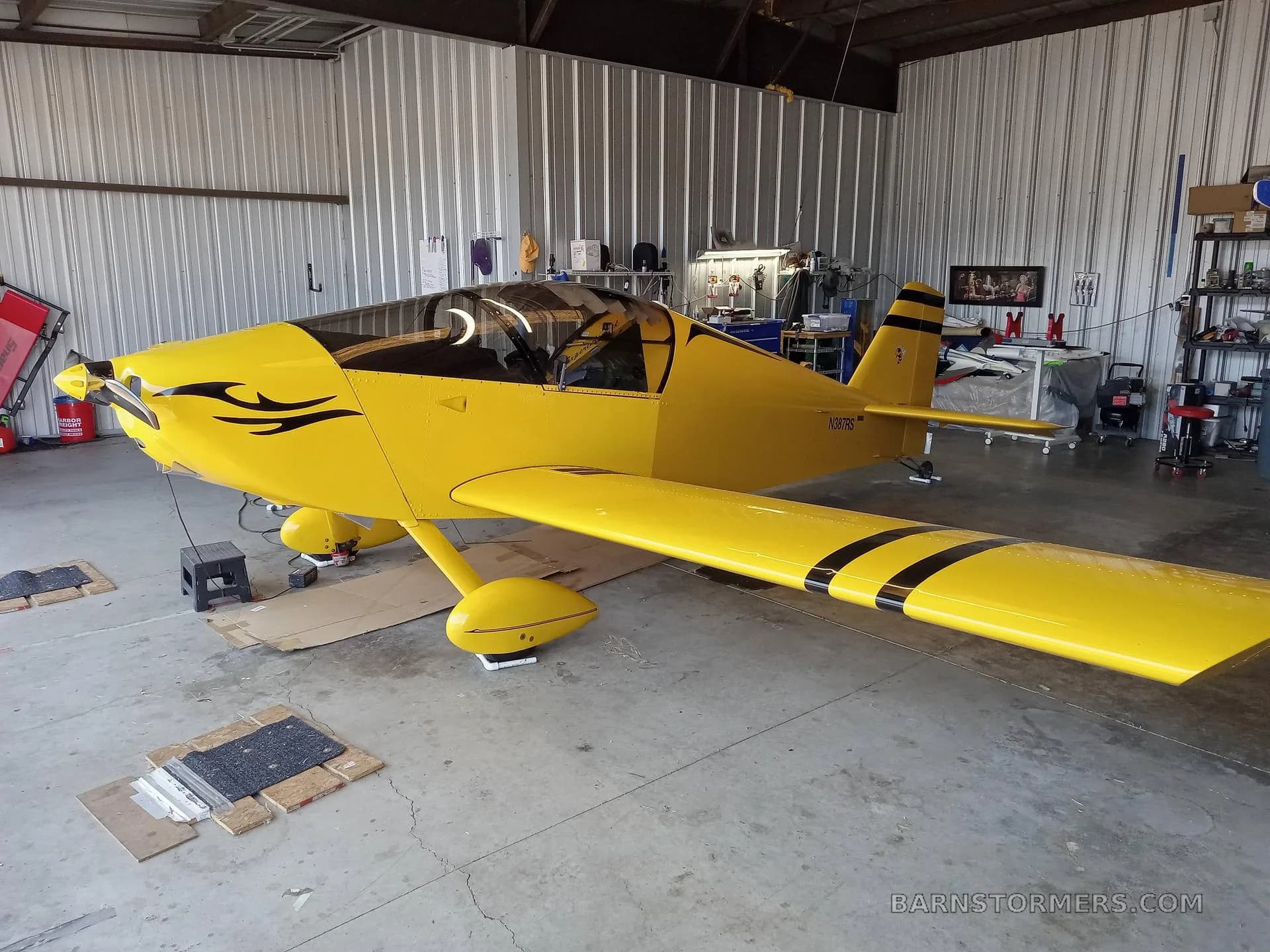 A bright yellow single-engine aircraft with black stripes and a black swirl decal on the nose, parked inside a hangar.