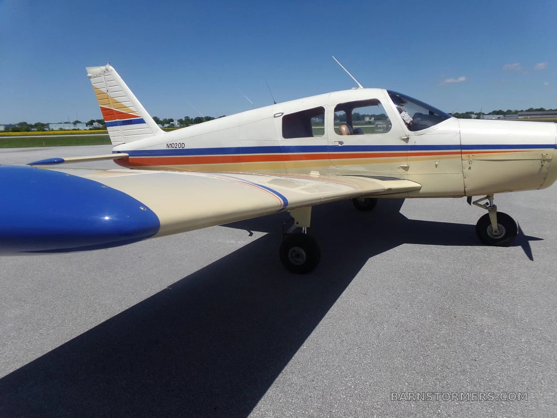 A close-up side view of a white and beige single-engine Piper aircraft with blue and orange stripes, showing the cockpit and wing.