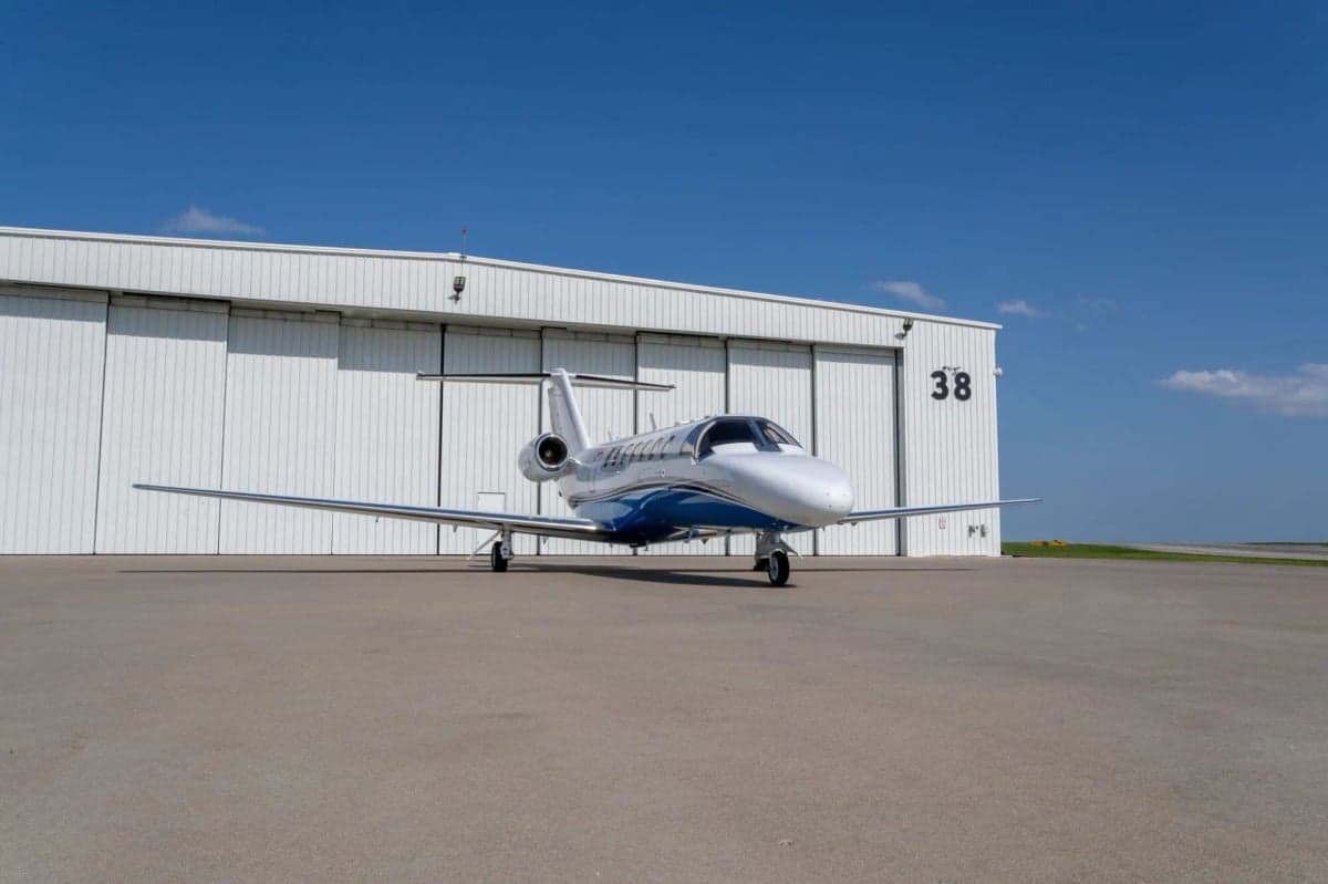 Front view of a white and blue jet aircraft parked on the tarmac in front of a hangar.