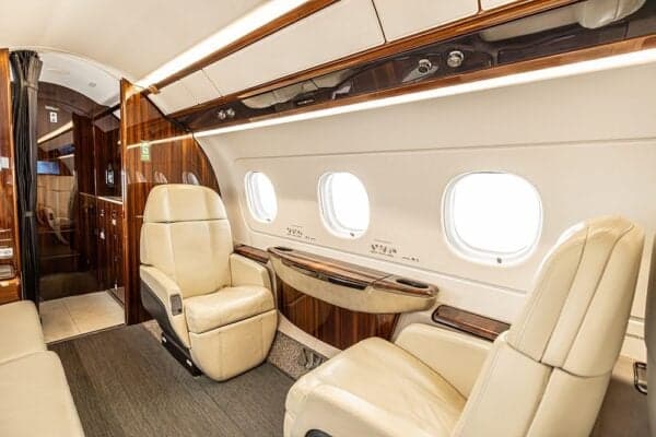 A view inside the Embraer Legacy 500 cabin, showing two cream-colored seats and a wooden side table.