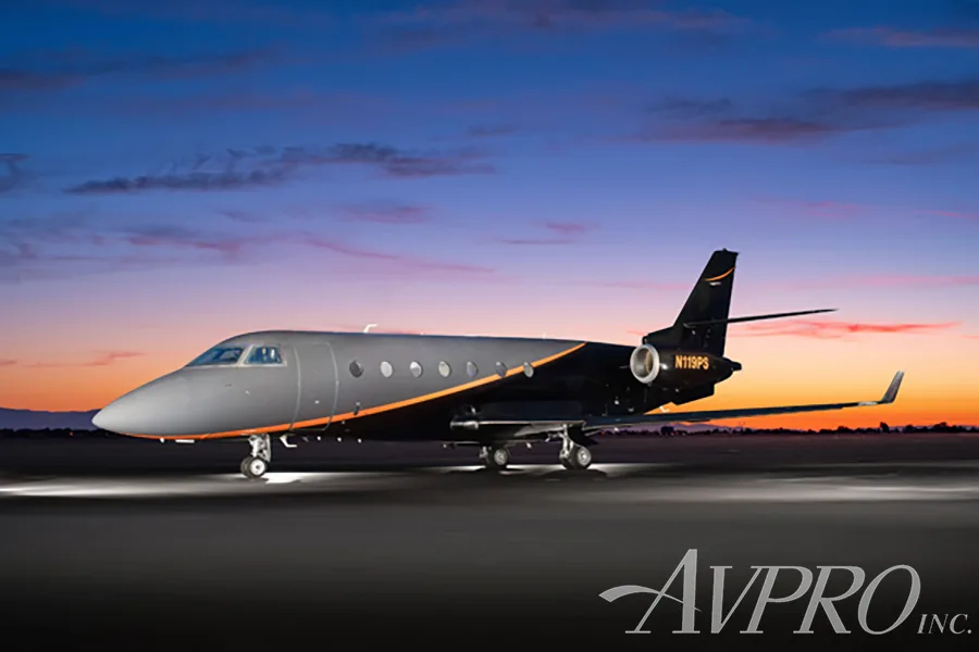 A Gulfstream G200 business jet, registration N119PS, with a dark gray and black paint scheme with orange accents, parked on an airfield at dusk.
