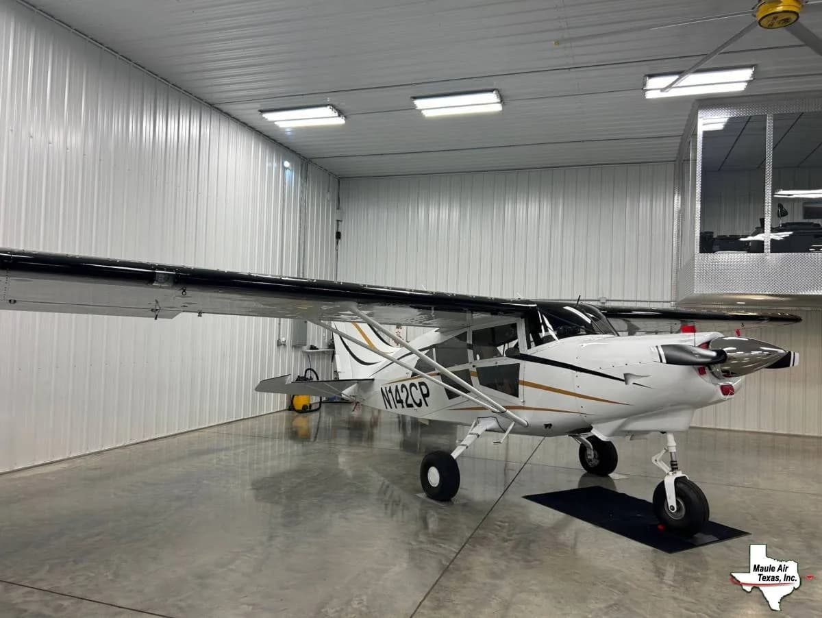 A white high-wing aircraft with black wings and gold stripes, featuring a nose wheel, sits in a hangar.